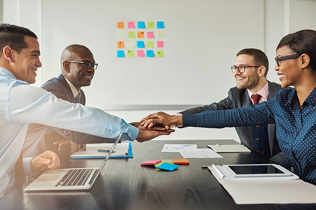 Image of business team reaching across the table stacking hands as they smile at each other.