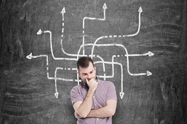 Man thinking in front of black chalk board with arrows pointing.