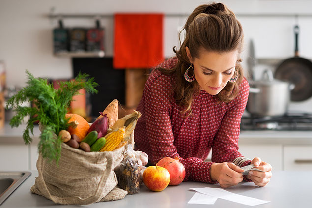 Woman reading shopping list, next to her on the kitchen counter is a burlap sac with a variety of fruits.