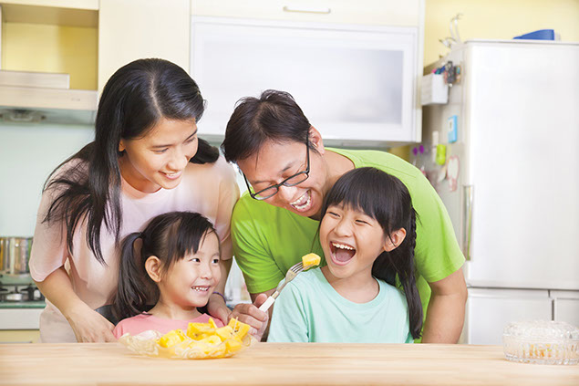 Image of laughing family as dad feeds daughter a piece of fruit.