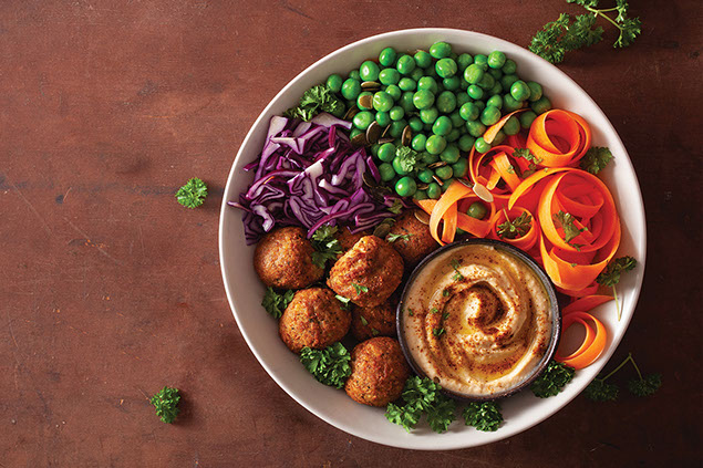 A bowl of falafel with hummus, green peas, carrots, and purple cabbage.