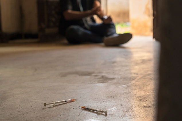 Image of needles on the ground and someone blurred out in the back injecting himself.